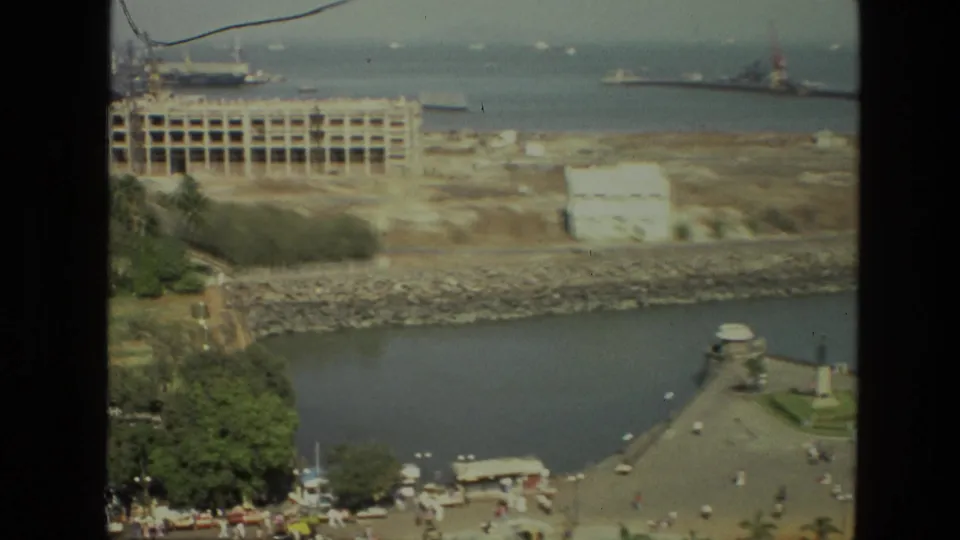 Elevated 1974 India waterfront view with pedestrians on a promenade, a small monument, rocky bank, unfinished structure, and distant ships.