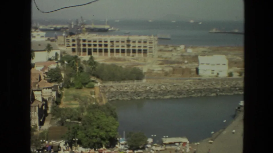 1974 India frame showing a harbor-like shoreline from above, with trees, low buildings, an unfinished structure, water, and ships offshore.