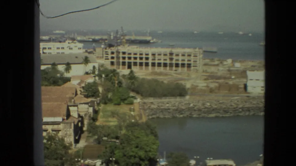 High-angle 1974 India home-movie view of a shoreline with trees, nearby buildings, a broad waterway, construction, and ships in the distance.