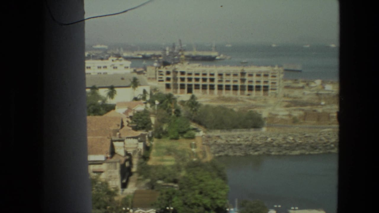 Coastal city view through a dark window frame with water, buildings, trees, and harbor structures beyond.