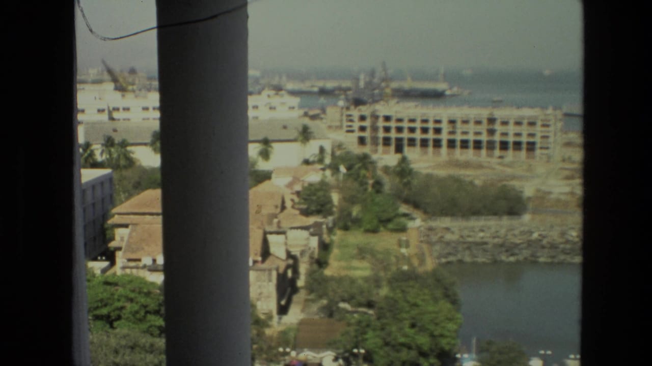 1974 India 8mm frame with a waterfront urban district, harbor cranes, ships, palms, and construction seen from an elevated interior viewpoint.