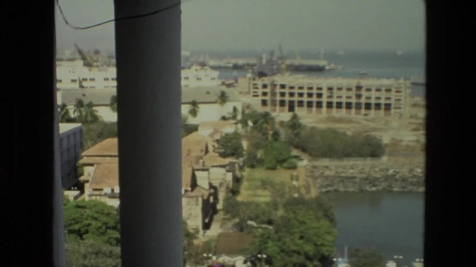 1974 India 8mm frame with a waterfront urban district, harbor cranes, ships, palms, and construction seen from an elevated interior viewpoint.