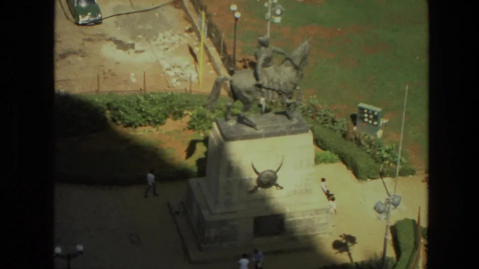 1974 India travel frame looking down on an equestrian statue, formal paths, low hedges, and pedestrians in a public plaza.