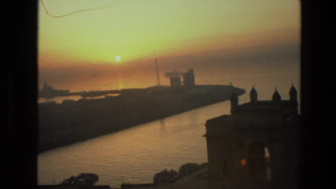 Warm sunset view over Mumbai harbor, with the Gateway of India in dark silhouette and distant waterfront structures across the water.