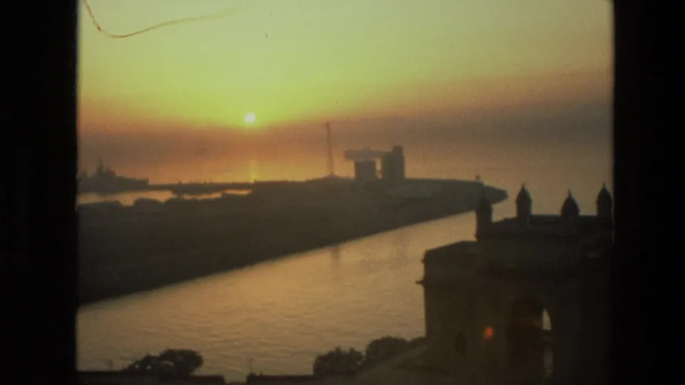 1974 home-movie frame of sunset over Mumbai harbor, with a dark waterfront silhouette and the Gateway of India cropped at the right.