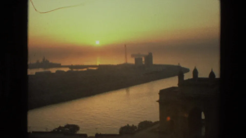 Late sequence frame showing the sun over Mumbai harbor, with warm haze, reflective water, and the Gateway of India partly visible in silhouette.