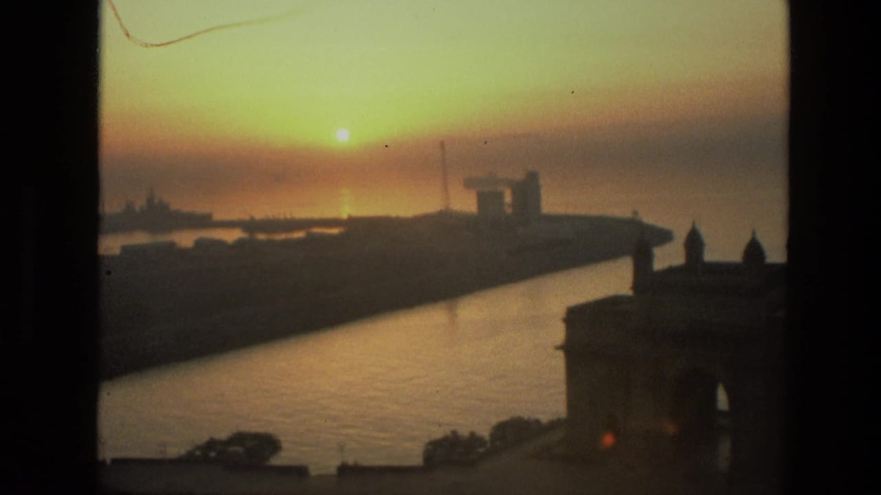 1974 8mm India harbor sunset with warm sky, reflective water, distant ships, and the Gateway of India silhouetted at right.
