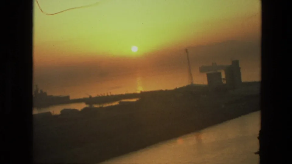 Soft 1974 India harbor sunset with orange reflection on the water, distant ship silhouettes, and shadowed waterfront architecture.