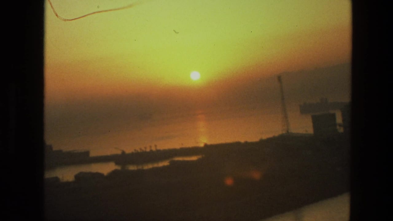 1974 India 8mm frame showing a hazy sun above a broad bay, with dark shoreline shapes and faint industrial silhouettes.