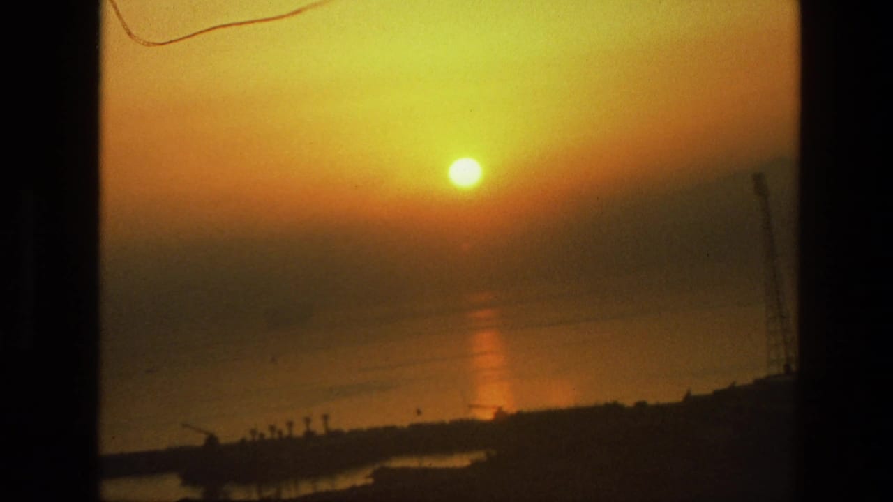 Orange sun over reflective water with a dark distant shoreline below.