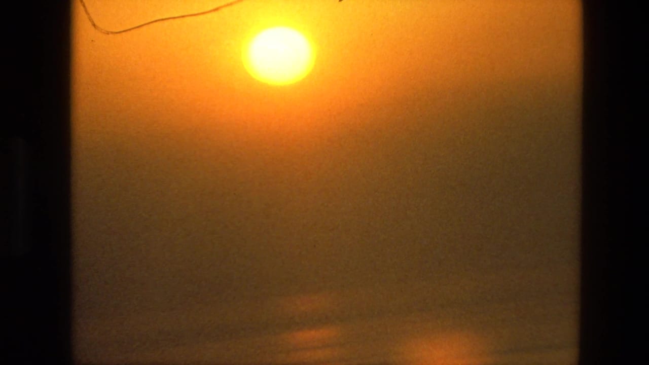 1974 8mm India frame showing an orange sun over hazy ocean water, partly framed by dark vertical edges.