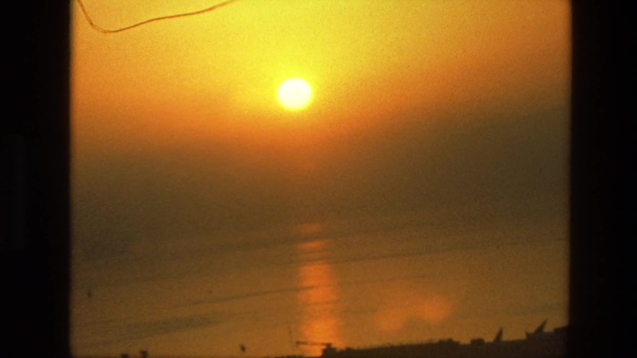1974 8mm India sunset view with the sun reflected on calm water and a low silhouetted shoreline at the bottom edge.