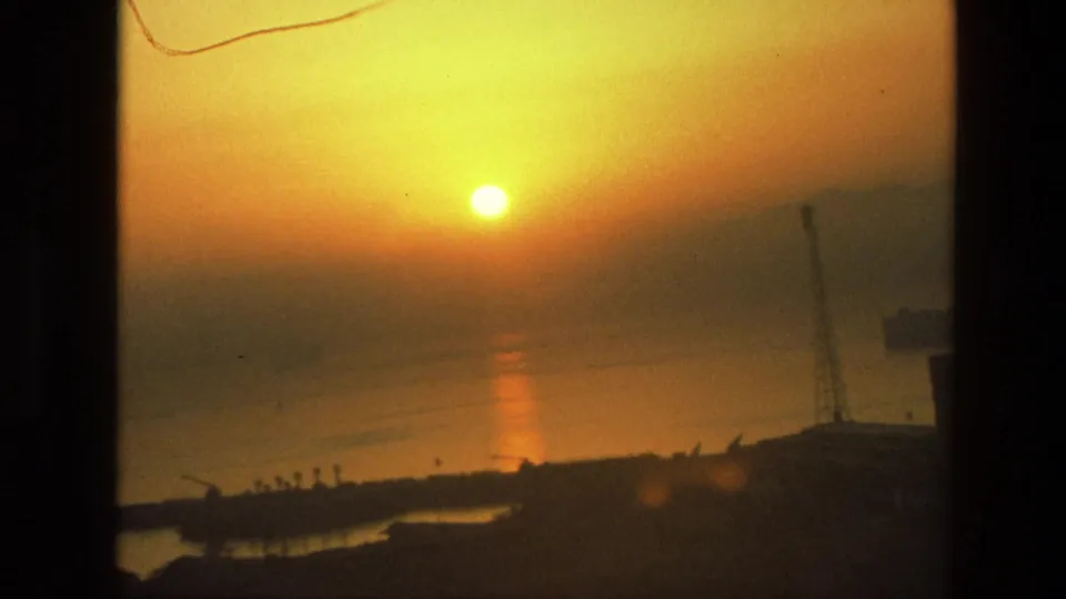 1974 8mm India frame showing a hazy orange sun, calm water reflection, and silhouetted waterfront structures with a crane.