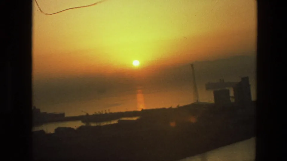1974 8mm India sunset frame with the sun near the horizon, reflected on water behind dark cranes and waterfront forms.