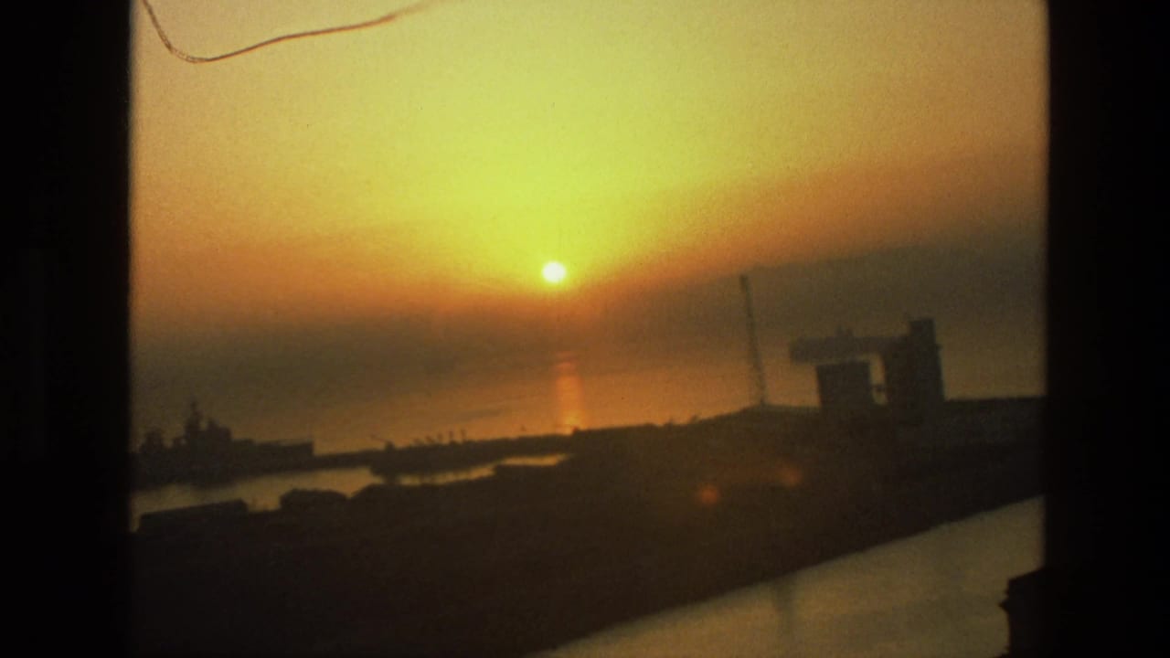 1974 India 8mm frame showing a low golden sun over coastal water, with silhouetted port structures and a dark foreground edge.