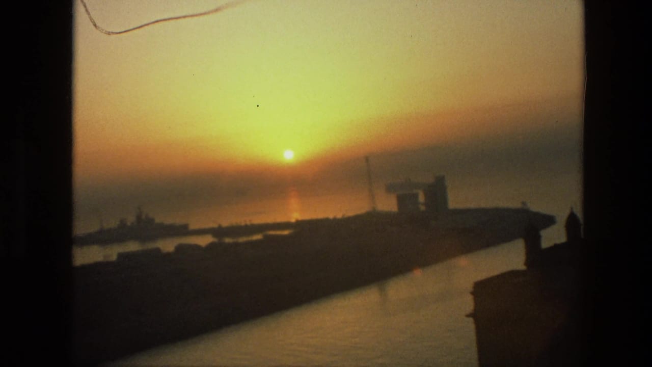 1974 India home-movie frame with sunset light on the water and shadowed harbor structures along the shoreline.