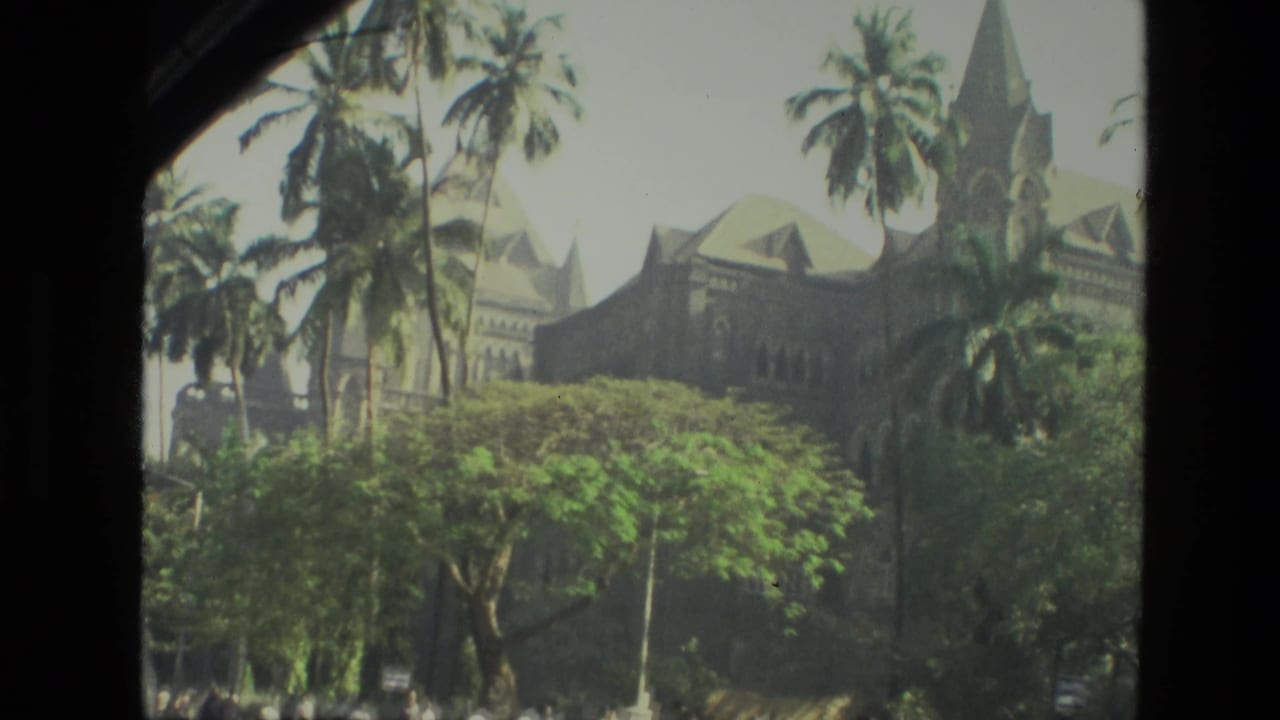1974 India 8mm frame showing a large stone building seen through palms and leafy trees from a visitor viewpoint.