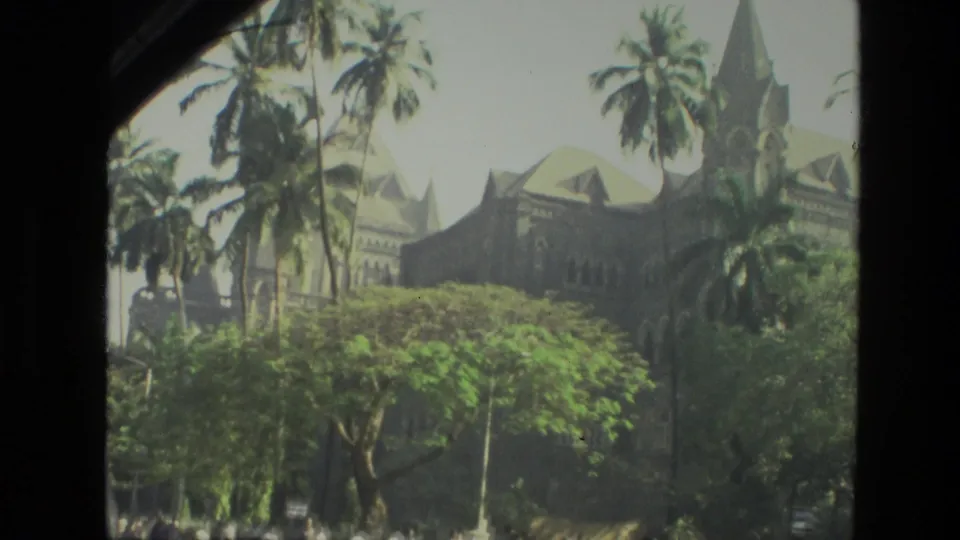 1974 India 8mm frame showing a large stone building seen through palms and leafy trees from a visitor viewpoint.