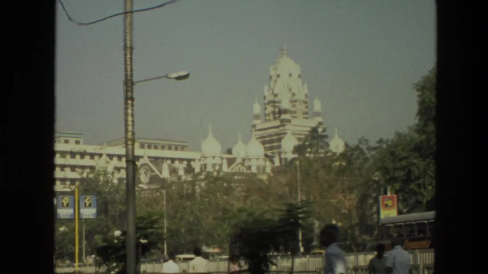 1974 India street view showing a domed building beyond lamps, buses, trees, and pedestrians in the foreground.