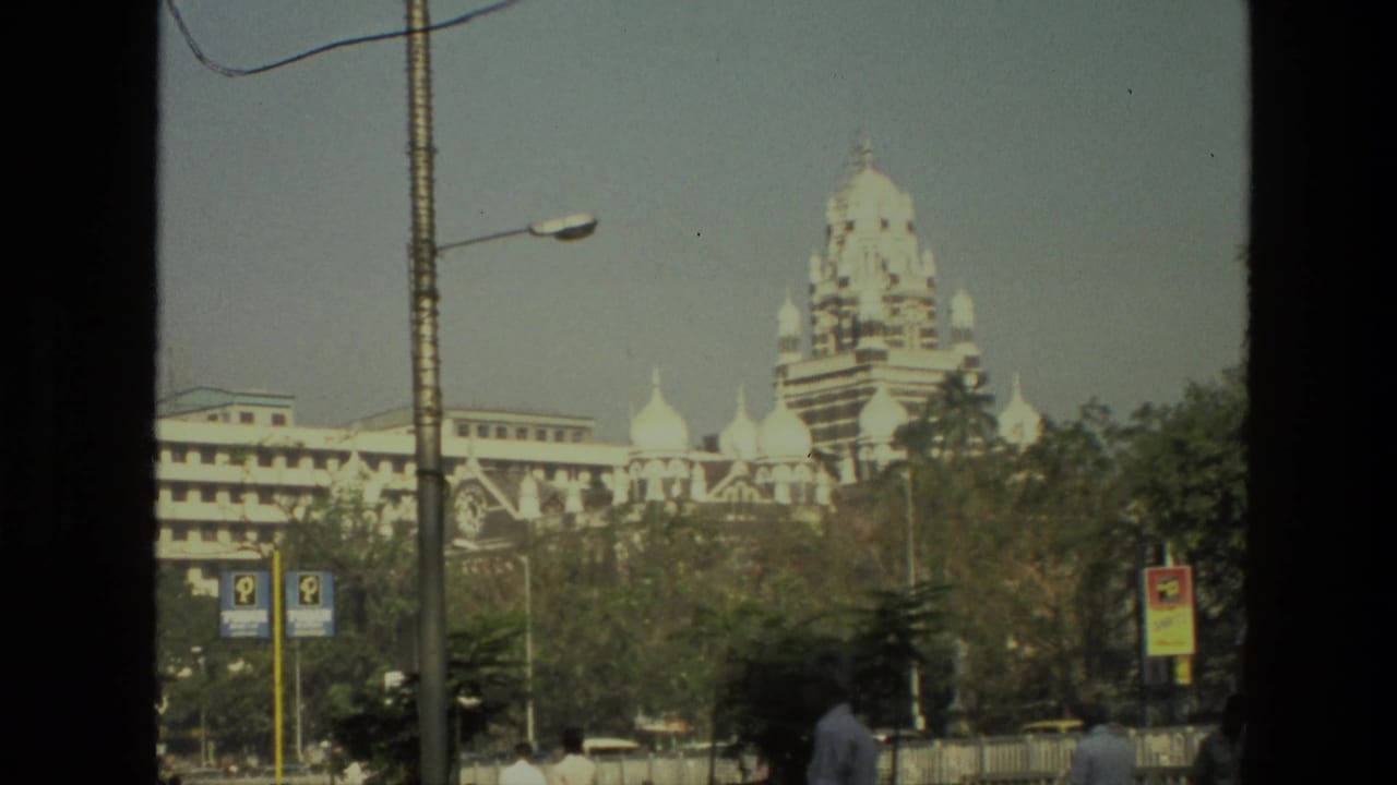 1974 India 8mm frame showing a large domed historic-looking building behind trees, with street lamps, parking signs, overhead wires, and pedestrians in the foreground.