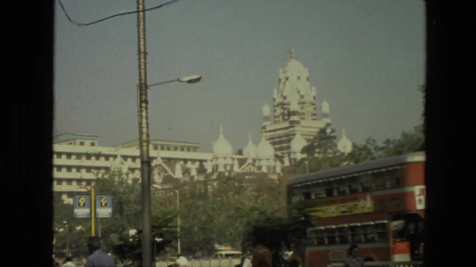 1974 India 8mm frame with a red double-decker bus crossing the foreground below a domed historic-looking building, trees, pedestrians, street lamps, and parking signs.
