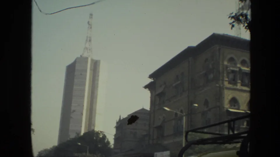 1974 India 8mm frame showing a tall modern building, an older arched facade, trees, street lighting, and a dark foreground edge from a vehicle or window.