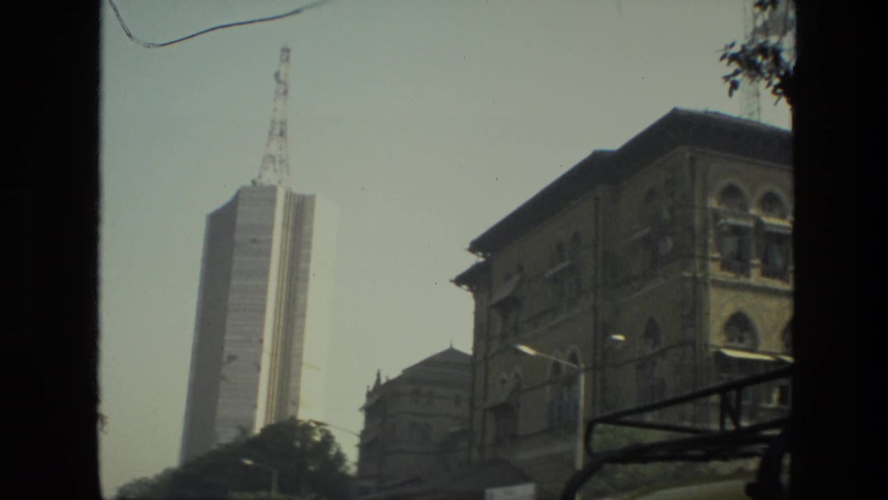 1974 India 8mm frame showing a tall communications tower rising near older urban buildings from a moving vehicle viewpoint.