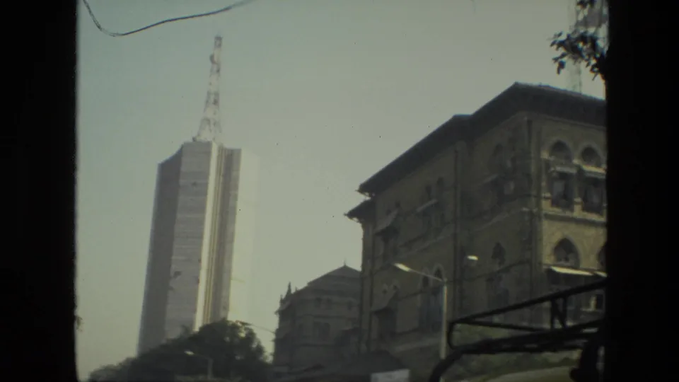1974 India 8mm frame showing a tall communications tower rising near older urban buildings from a moving vehicle viewpoint.