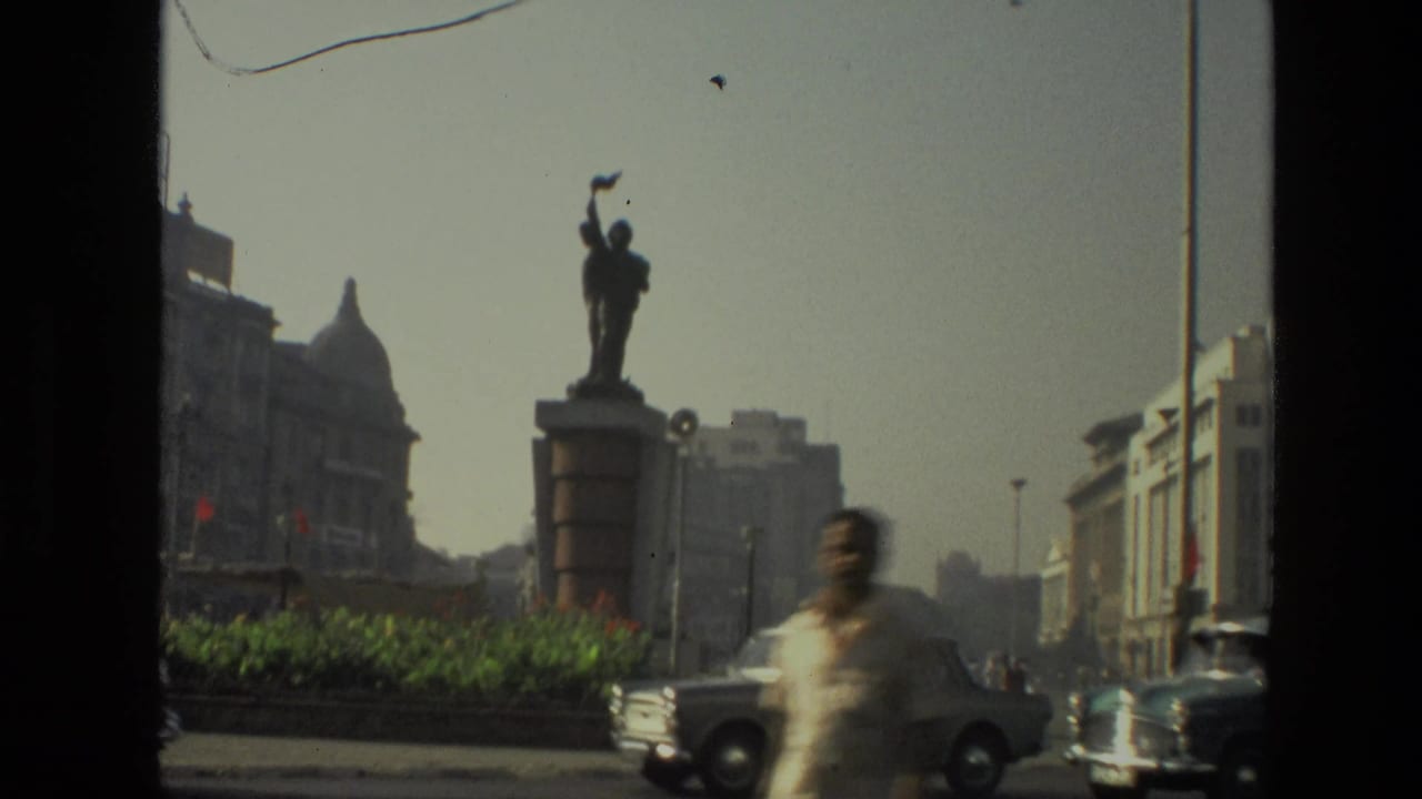 1974 India street frame with a statue on a pedestal, passing cars, a blurred pedestrian, and historic civic buildings in the background.