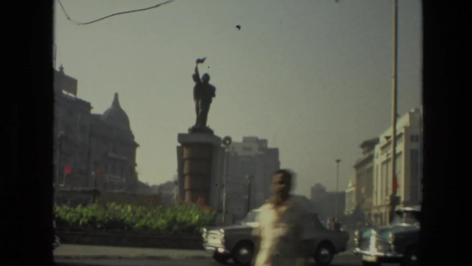1974 India street frame with a statue on a pedestal, passing cars, a blurred pedestrian, and historic civic buildings in the background.