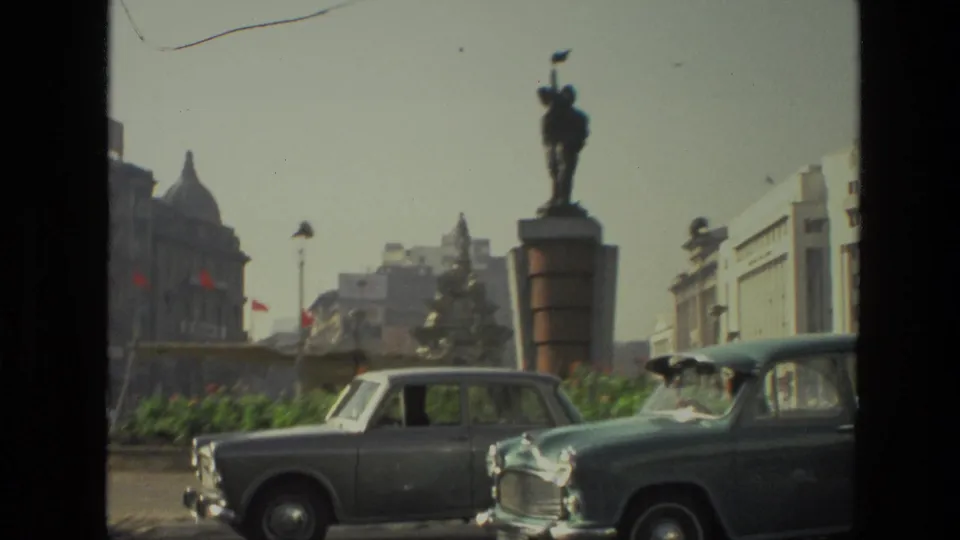 1974 India 8mm frame showing classic cars near a statue, planted median, streetlights, and historic buildings beyond.