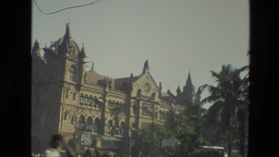 1974 India frame showing an ornate historic stone building, palm trees, street vegetation, and a blurred pedestrian in the foreground.