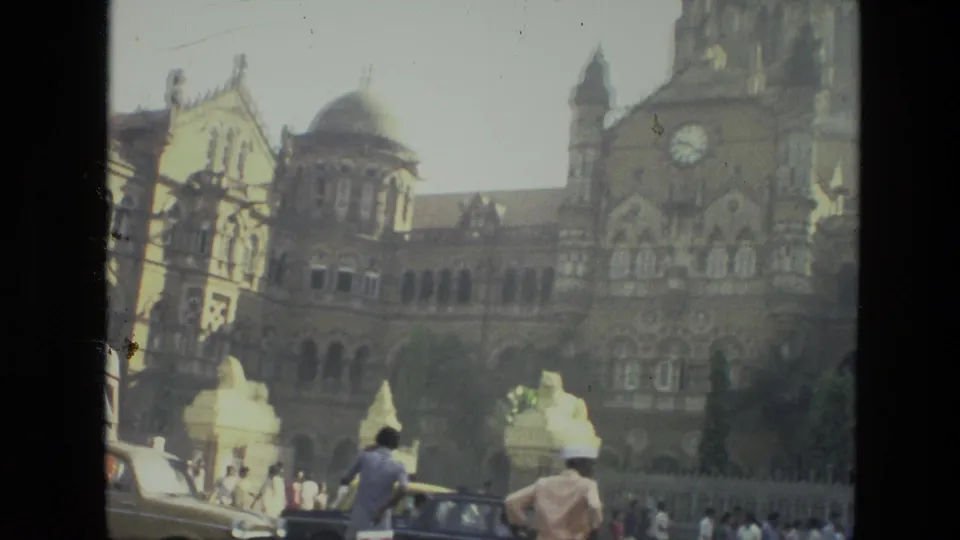 1974 Mumbai street view of the terminus clock tower and dome, with cars and small pedestrian figures near the entrance area.