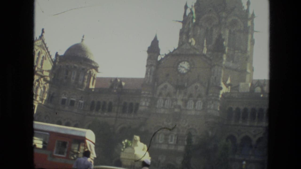 1974 India 8mm frame showing pedestrians and a red bus near a large ornate historic building with domes and a clock tower.