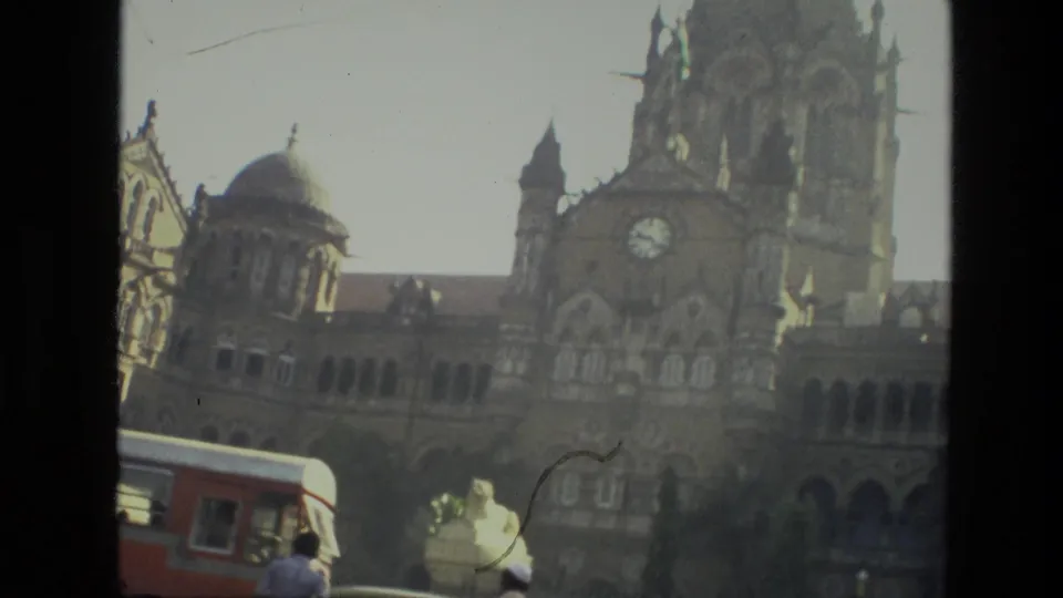 1974 India 8mm frame showing pedestrians and a red bus near a large ornate historic building with domes and a clock tower.