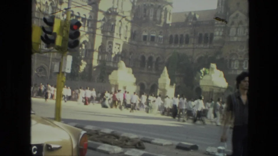 1974 India street scene with pedestrians, traffic lights, cars, and a large ornate historic building in the background.