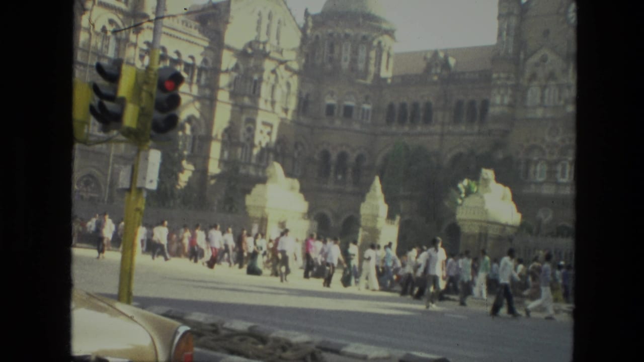 1974 India 8mm frame showing pedestrians crossing near an ornate colonial-style building, with a traffic signal and car edge in the foreground.