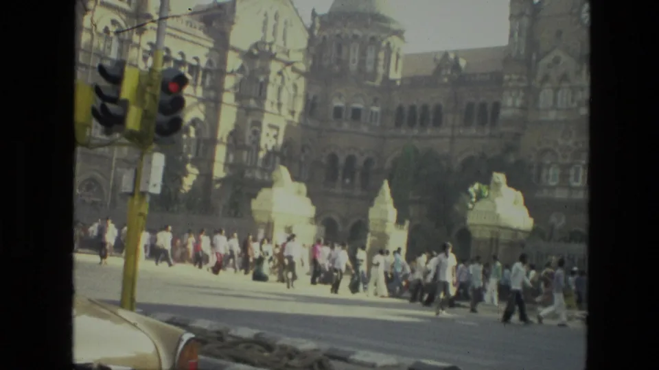 1974 India 8mm frame showing pedestrians crossing near an ornate colonial-style building, with a traffic signal and car edge in the foreground.