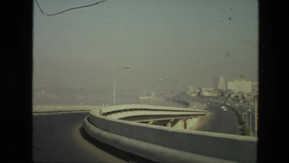 1974 India 8mm frame showing a curved elevated roadway, streetlights, distant buildings, and haze from a moving vehicle viewpoint.