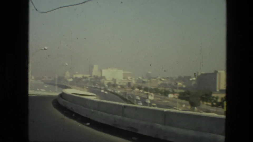 1974 India travel frame with a concrete roadway barrier in the foreground, distant traffic, low skyline, and bright haze.