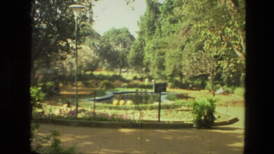 1974 India 8mm frame showing a bright garden scene with paths, trees, lampposts, railings, and a low fountain area.