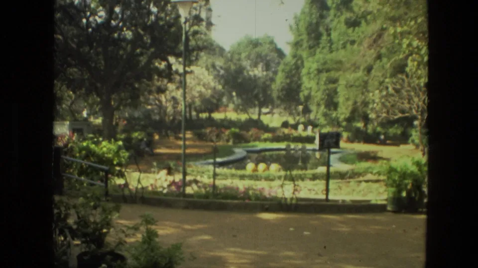 1974 India home-movie frame of a landscaped park with a curved fountain edge, mature trees, railings, lampposts, and planting beds.