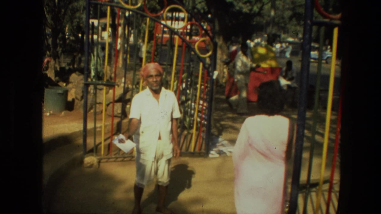 In 1974 India home-movie footage, a man in white clothing and red turban stands by a colorful metal park gate near a woman in a pale sari.