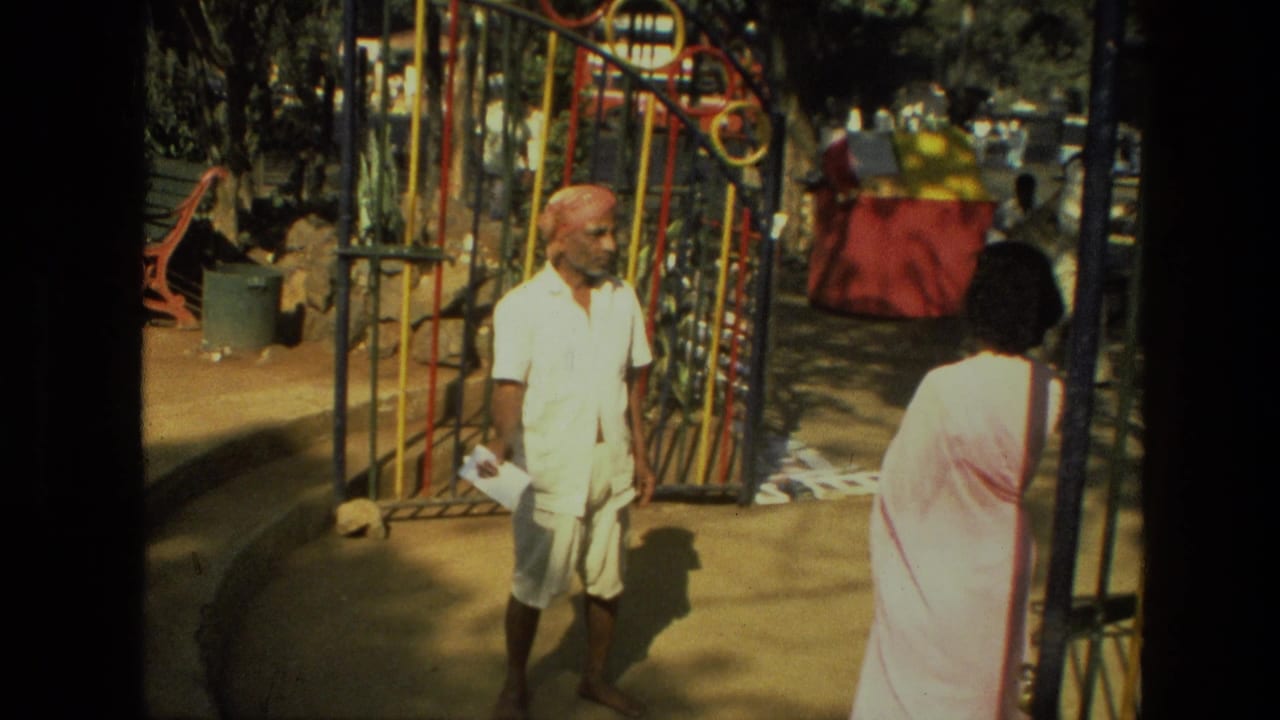 The man in white clothing and red turban holds paper near the park gate, with the sari-clad woman partly visible at frame right.