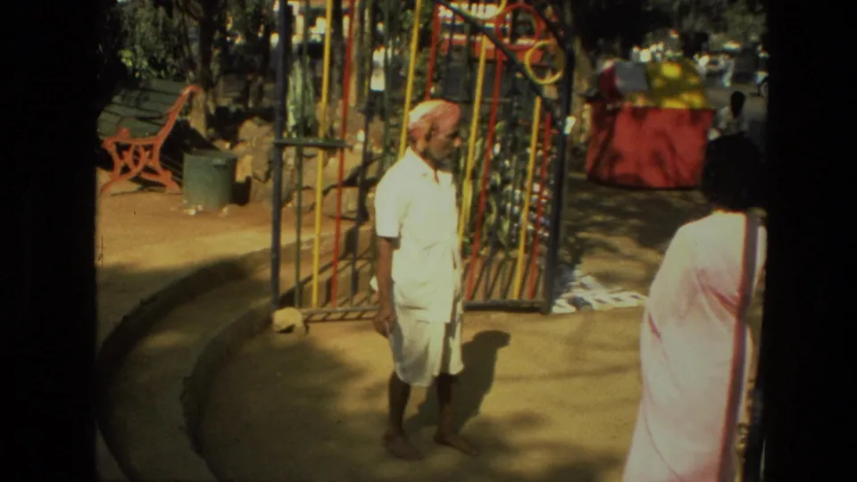 A man in white clothing and red turban pauses beside an open metal park gate, with a woman in a pale sari seen from behind.