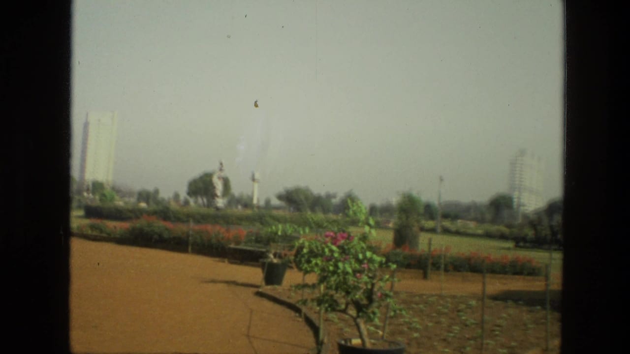 1974 India park view with planted flower beds, potted shrubs, open lawn, lamp posts, and distant high-rise buildings.