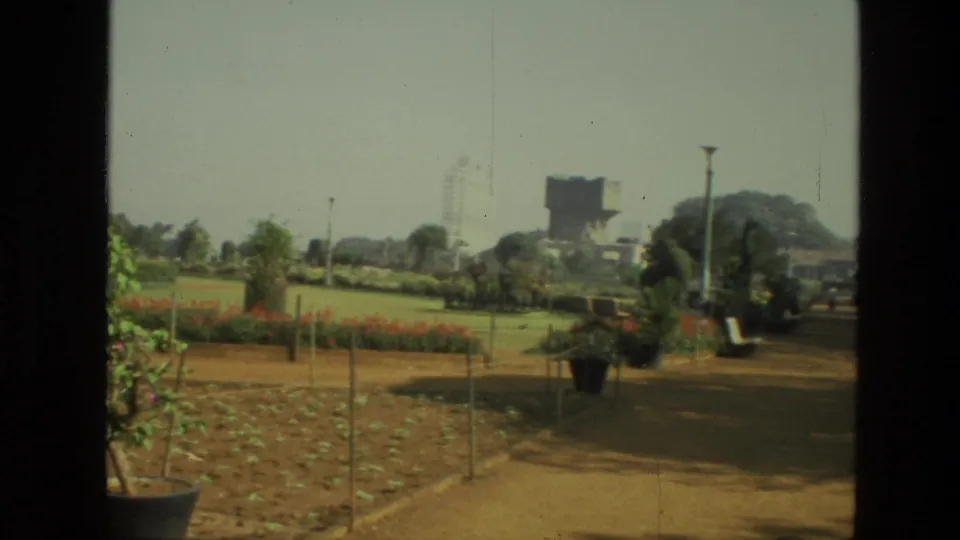 1974 India home-movie frame with a curving dirt path, planted beds, potted greenery, open lawn, trees, and distant high-rises.