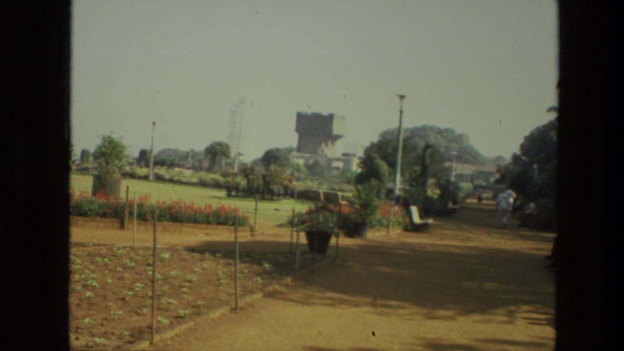 Dirt path through a landscaped park with flower beds, potted plants, trees, and people in the distance.