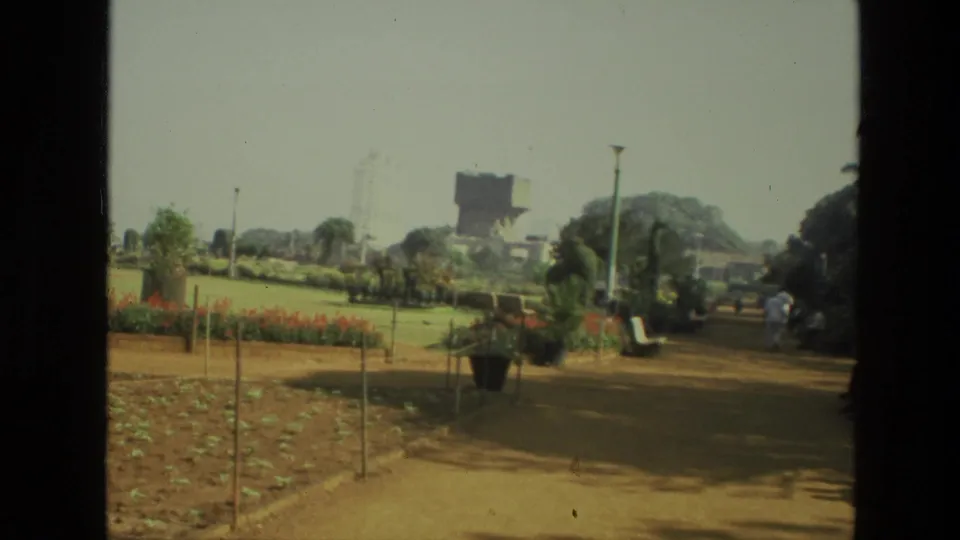 1974 India 8mm view of a landscaped urban park path with plant beds, trees, benches, and distant visitors.