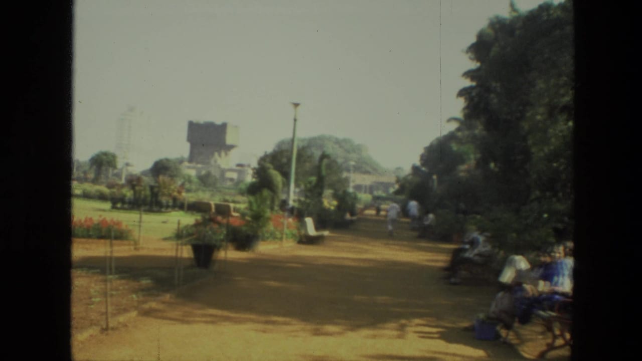 1974 India park view with a dirt path, manicured planting, benches, trees, and visitors sitting or walking nearby.